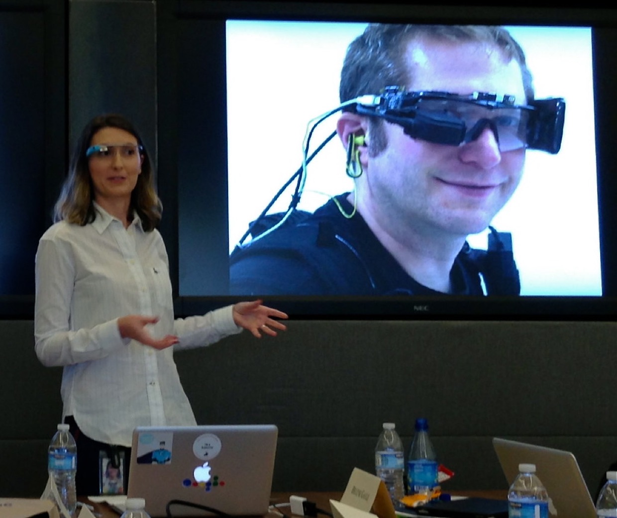 A woman wearing Google Glass presenting in a meeting room, with a projected photograph behind her of an earlier head-mounted wearable computer.